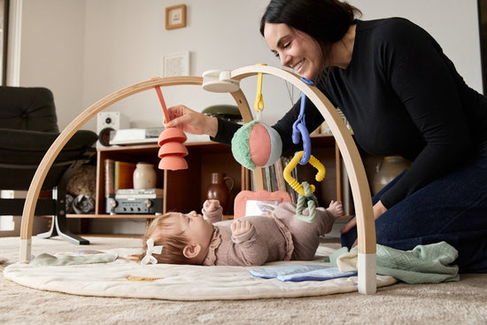 Baby playing with a wooden arch toy on a rug in a room with mom