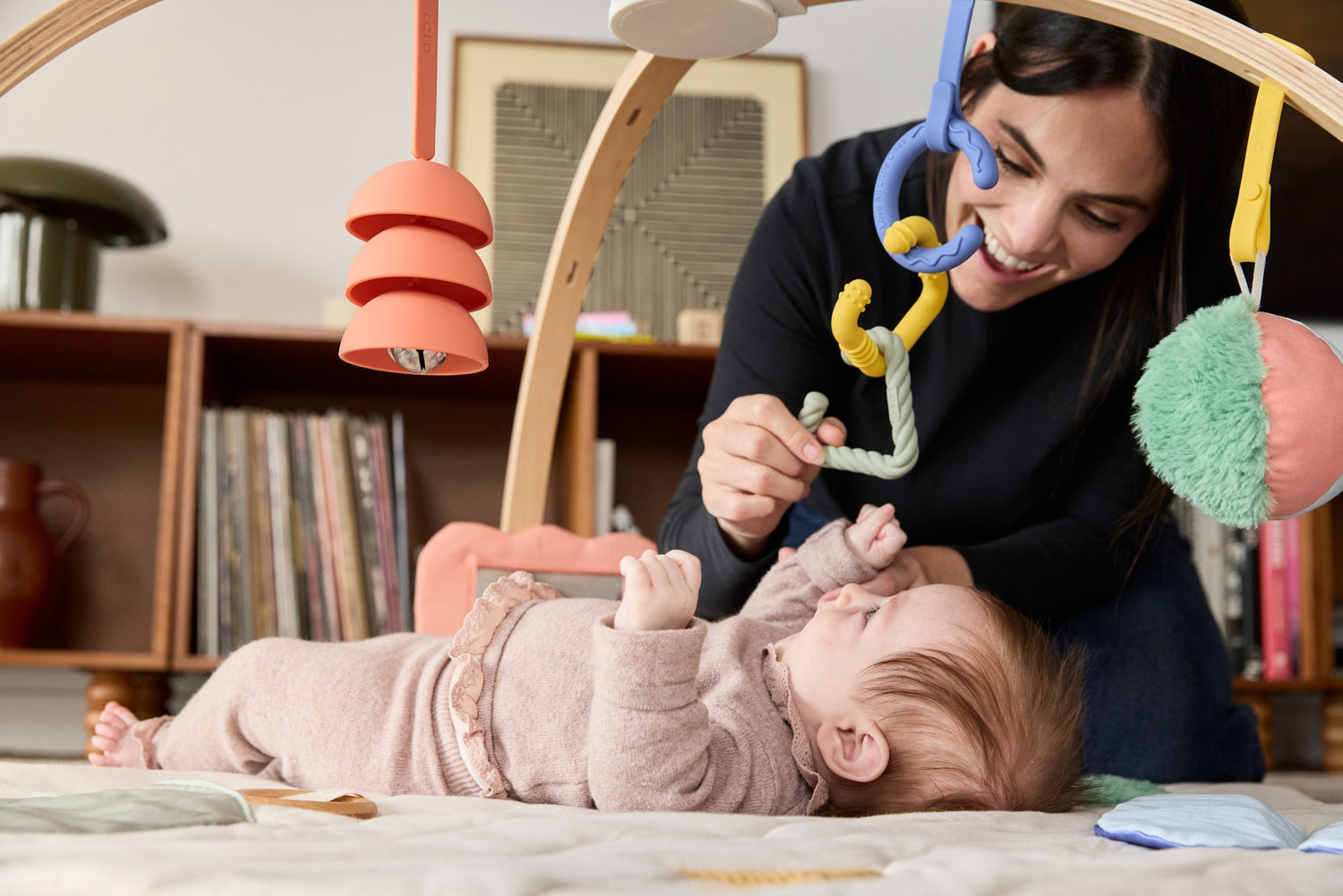 Woman playing with a baby on a play mat with colorful toys in a room.